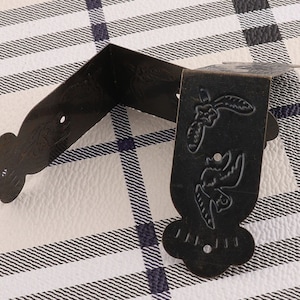 May include: Two black metal corner brackets with decorative floral and bird designs. The brackets are on a white and black checkered surface.