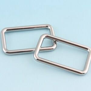 May include: Two silver metal rectangular rings, one slightly larger than the other, are stacked on top of each other. The rings are smooth and shiny.