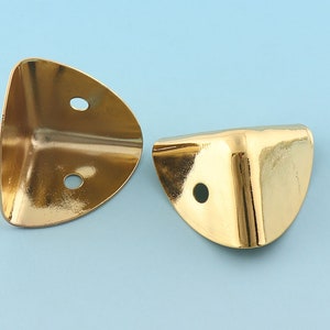 May include: Two gold metal corner protectors with rounded edges and two holes each. The protectors are designed to protect furniture corners from damage.