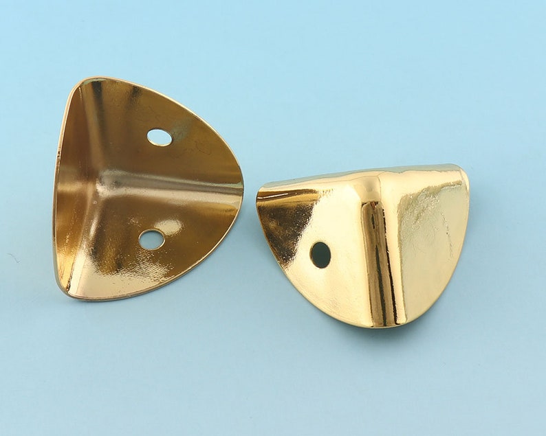 May include: Two gold metal corner protectors with rounded edges and two holes each. The protectors are designed to protect furniture corners from damage.