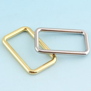 Puede incluir: Dos anillos metálicos, uno dorado y otro plateado, están apilados uno encima del otro. Los anillos son de forma rectangular y tienen un acabado liso y pulido.