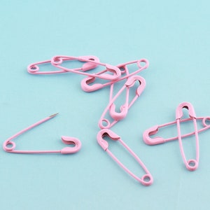 Pu&ograve; includere: Un gruppo di spille da balia rosa su uno sfondo blu chiaro. Le spille sono disposte in un mucchio sciolto, con alcune che si sovrappongono.