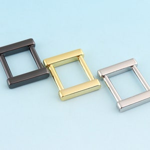 Puede incluir: Tres anillos en D de metal en negro, dorado y plateado. Los anillos son todos del mismo tamaño y forma, y están dispuestos en una fila de izquierda a derecha.