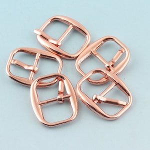 Puede incluir: Cinco hebillas de cinturón de metal de color oro rosa. Las hebillas son rectangulares, con un acabado liso y una barra central.