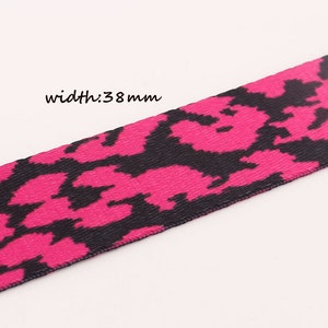 May include: Pink and black patterned ribbon with a width of 38 mm. The pattern is a repeating design of black shapes on a pink background.