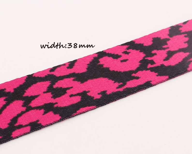 May include: Pink and black patterned ribbon with a width of 38 mm. The pattern is a repeating design of black shapes on a pink background.