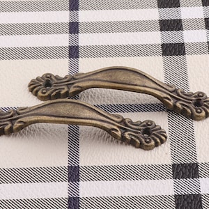 Puede incluir: Dos tiradores de armario de estilo antiguo con diseños ornamentados. Los tiradores son de metal y tienen un acabado bronce.