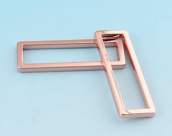 May include: Two rose gold metal belt buckle loops. The loops are rectangular and have a smooth, polished finish.
