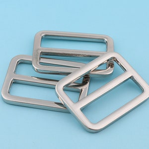 Puede incluir: Cuatro hebillas deslizantes de metal plateado. Las hebillas son rectangulares con un borde redondeado y una abertura central. Las hebillas están apiladas una encima de la otra.