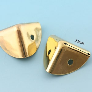 May include: Two gold metal corner protectors, each with a rounded edge and a hole in the center. The corner protector on the right has a measurement of 25mm.