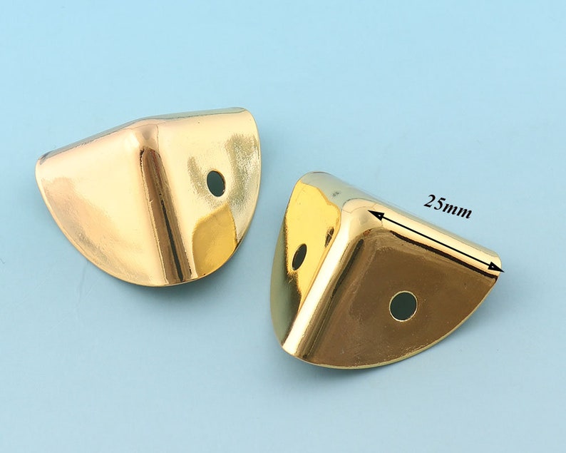 May include: Two gold metal corner protectors, each with a rounded edge and a hole in the center. The corner protector on the right has a measurement of 25mm.