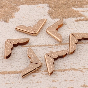 May include: Six rose gold metal corner protectors with an ornate, curved design. The protectors are arranged on a light-coloured, textured surface. Each corner piece has a decorative, etched pattern.