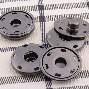 May include: A set of five black metal snap buttons. The buttons have a round shape with a center hole and a raised rim. The buttons are designed for sewing onto fabric.