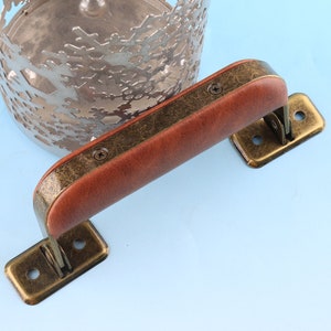 May include: A brown leather handle with brass accents and screws. The handle is attached to a brass base with two screws. The handle is designed to be used on a piece of furniture.