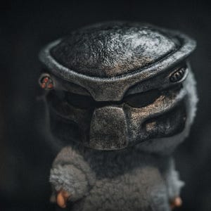 May include: A plush toy with a Predator mask in metallic grey, with red accents. The toy has a grey, furry body and small orange hands, set against a dark background. The mask is detailed with textured surfaces.