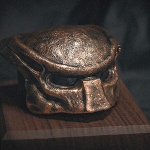 May include: A bronze Predator mask, displayed on a wooden plinth. The mask showcases intricate detailing, including the distinctive mandibles and eye openings. The metallic finish has a weathered look, implying a battle-hardened appearance. The wooden base offers a contrasting, natural element.
