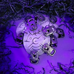 Puede incluir: Un conjunto de cierres metálicos plateados dispuestos sobre una superficie circular clara con diseños grabados. Los cierres son rectangulares con un diseño de bisagra. El fondo es morado oscuro con papel triturado.