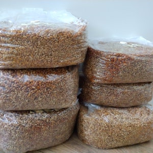 May include: Six clear plastic bags filled with brown wheat berries are stacked on top of each other. The bags are sealed and ready to be used.