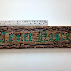 Pode incluir: Uma placa retangular de madeira com acabamento tingido de castanho escuro e fundo verde rebaixado. O texto "Temet Nosce" está esculpido numa fonte gótica. Uma régua metálica colocada acima mostra um comprimento de cerca de 30 cm.