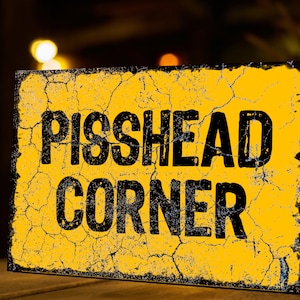 May include: A yellow sign with black text that reads "PISSHEAD CORNER". The sign is weathered and has cracks in the paint.