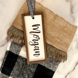 May include: A black and white buffalo plaid stocking with a brown wooden tag that says "Morgan" in cursive. The stocking has a brown burlap cuff with a faux fur trim.