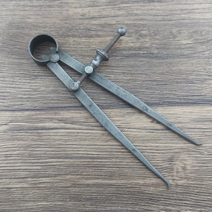 May include: A silver metal compass with a circular handle and a pointed tip. The compass is on a wooden surface.