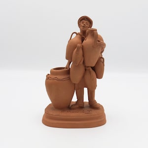 Puede incluir: Escultura de terracota de un hombre que lleva varias jarras y un jarrón grande. La figura viste ropa sencilla y un sombrero. La escultura está sobre una base redonda con texto.