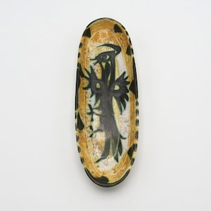 Può includere: Un piatto ovale in ceramica con un design nero, verde e giallo. Il design presenta un uccello stilizzato con le ali spiegate.