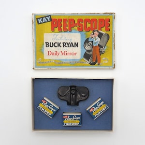 May include: A vintage cardboard box with a blue and white interior. The box contains a black plastic viewer and three film strips. The box lid features a colorful illustration of a boy holding a camera and the text "KAY PEEP-SCOPE Featuring BUCK RYAN of the Daily Mirror".