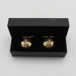 May include: Gold-coloured cufflinks with an emblem design, displayed in a black box. The round cufflinks have a shiny finish, suitable for formal occasions. Each cufflink features a detailed emblem.