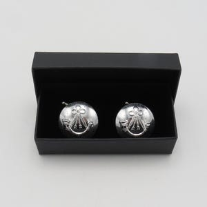 Puede incluir: Un par de gemelos plateados con un diseño de emblema detallado, presentados en una caja negra abierta. Los gemelos tienen forma circular con un patrón decorativo. La caja es rectangular y tiene un acabado mate.