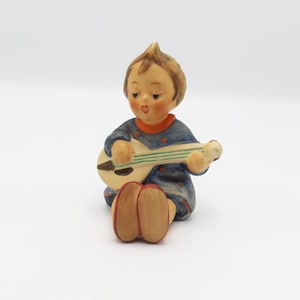 May include: A ceramic figurine of a seated child playing a small guitar. The child has brown hair, rosy cheeks, and is wearing a blue outfit with red accents. The guitar is cream-colored with green stripes. The figurine is on a white background.