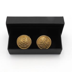 Może przedstawiać: Dwa złote spinki do mankietów z kanadyjskim godłem przedstawiającym liść klonu, skrzyżowane karabiny i napis "CANADA". Spinki do mankietów są prezentowane w czarnym pudełku.