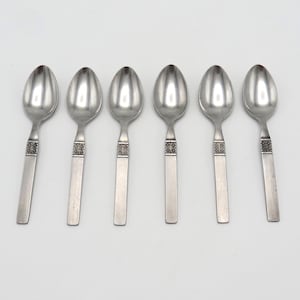 Può includere: Sei cucchiai in acciaio inossidabile su sfondo bianco. Ogni cucchiaio ha una forma arrotondata e un manico rettangolare con un motivo decorativo. I cucchiai sono distanziati uniformemente e riflettono la luce.