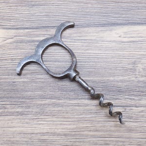 Op de afbeelding: Een antieke metalen kurkentrekker. Het heeft een cirkelvormige handgreep met gebogen elementen en een spiraalvormige worm. Het metaal heeft een versleten, verouderd uiterlijk.
