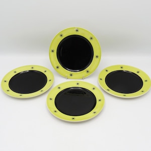 May include: Four ceramic plates with a black centre and a light green border. The border features small starburst designs. The plates are arranged on a white surface.