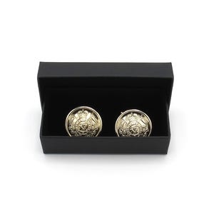 May include: A pair of gold-coloured cufflinks with a detailed emblem design, presented in an open black box. The circular cufflinks have a shiny finish, set against a white background.