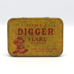 May include: A rectangular, vintage tobacco tin with a yellow and distressed finish. The tin features red text that reads "PLAYER'S DIGGER FLAKE TOBACCO" and an illustration of a man smoking a pipe. The text "JOHN PLAYER & SONS, NOTTINGHAM" is also visible.