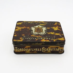 May include: A vintage tin box for Tortoiseshell Cigarettes. The box is brown and yellow with a tortoise shell pattern and a gold emblem of a turtle. The text "Tortoiseshell Cigarettes" is printed on the bottom of the box.