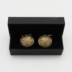 Może przedstawiać: Para złotych spinek do mankietów z misternym wzorem herbu, zapakowanych w czarne pudełko. Okrągłe spinki do mankietów mają błyszczące wykończenie i nadają się do formalnych okazji.