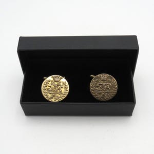 May include: Gold-coloured cufflinks with an emblem design, displayed in a black box. The circular cufflinks feature a crown and laurel wreath motif. The box is open, revealing the cufflinks.