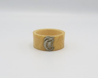 May include: A cream-colored napkin ring with a silver-colored letter "T" in a decorative font.