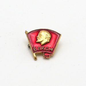 May include: A red and gold enamel pin featuring a profile of a man and the text "ВЛКСМ" in Cyrillic letters.