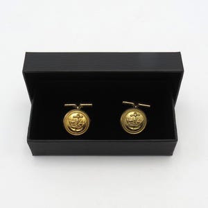 Può includere: Un paio di gemelli color oro con un design dettagliato di corona, croce e nave, presentati in una scatola nera. I gemelli hanno una forma circolare classica e una finitura lucida, adatti per occasioni formali.