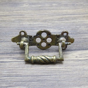 Puede incluir: Tirador de cajón de latón antiguo con un diseño decorativo. El tirador presenta una sección central con cinco recortes circulares y un asa retorcida. Las placas de montaje tienen detalles ornamentados. Herrajes para la restauración de muebles.