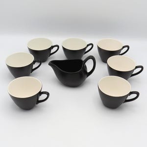 Peut inclure: Un ensemble de tasses à thé noires et crème et une crémière. Les tasses ont un extérieur noir et un intérieur crème. La crémière est noire. L'ensemble est disposé sur une surface blanche.