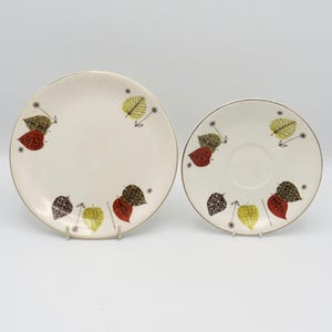 May include: A cream-coloured ceramic plate and saucer set. The set has a gold rim and is decorated with colourful leaf designs in red, yellow, and brown. The plate is larger than the saucer.
