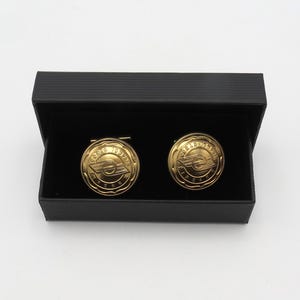 May include: A pair of gold-colored cufflinks with a circular design, presented in a black box. The cufflinks feature an emblem with text and a stylized wing design. The box is open, revealing the cufflinks against a black interior.