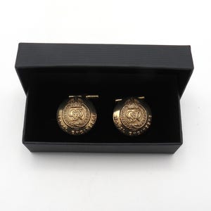 Puede incluir: Un par de gemelos dorados con las palabras "ROYAL ENGINEERS" y un diseño de escudo, presentados en una caja negra. Los gemelos tienen forma circular con un emblema detallado.