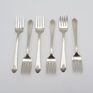 May include: Six silver-plated forks with ornate handles. The forks are arranged in a row on a white background.